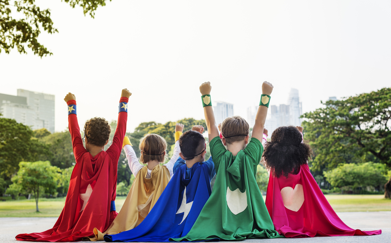 Back view of five young children sitting on the ground wearing superhero capes of various primary colors